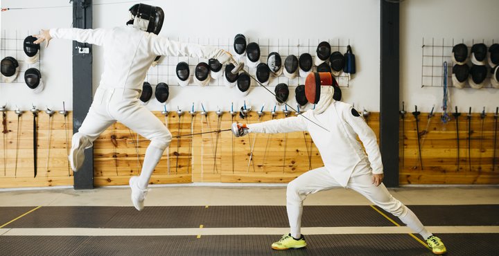 Unrecognizable students fencers in protective uniform and with swords dueling during training in gym