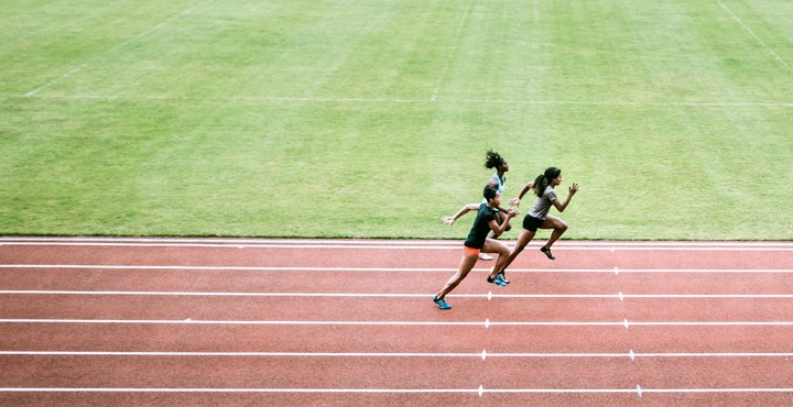 Young adult women sprint down the track, viewed from high in the stadium seating.  Wide high angle view.  Training for track and field competition.